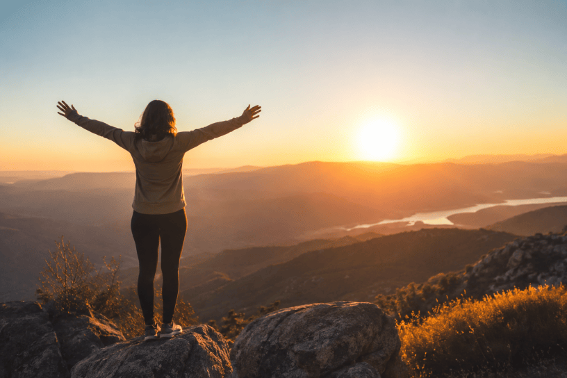 Persona in piedi su una vetta montana al tramonto con le braccia aperte verso l’orizzonte, simbolo di resilienza, libertà interiore e forza mentale.