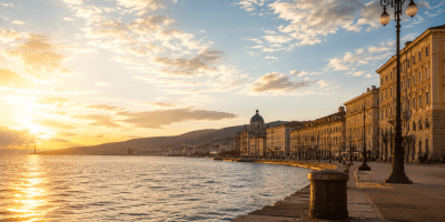 Panorama del lungomare di Trieste al tramonto, con il sole che si riflette sul mare Adriatico, edifici storici illuminati dalla luce dorata e cielo attraversato da nuvole estive.