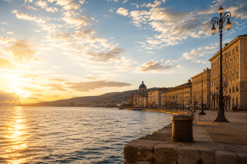 Panorama del lungomare di Trieste al tramonto, con il sole che si riflette sul mare Adriatico, edifici storici illuminati dalla luce dorata e cielo attraversato da nuvole estive.