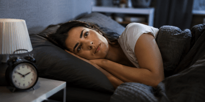 Persona sdraiata nel letto al mattino con espressione stanca e affaticata, mentre la luce dell'alba entra dalla finestra della camera da letto.