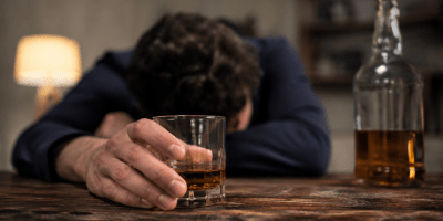 Uomo adulto seduto da solo davanti a un bicchiere di alcol in un ambiente domestico poco illuminato, con espressione pensierosa e preoccupata. L'immagine rappresenta le conseguenze psicologiche e sociali della dipendenza da alcol.