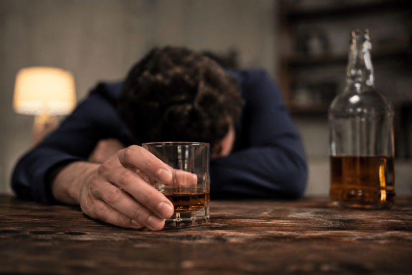 Uomo adulto seduto da solo davanti a un bicchiere di alcol in un ambiente domestico poco illuminato, con espressione pensierosa e preoccupata. L'immagine rappresenta le conseguenze psicologiche e sociali della dipendenza da alcol.