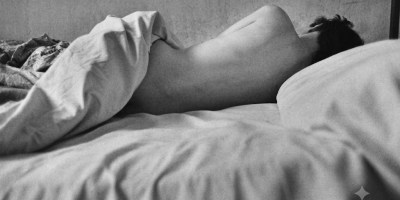 Fotografia artistica in bianco e nero di una figura femminile distesa su un letto, avvolta dalle lenzuola, in un'atmosfera intima, contemplativa e poetica.