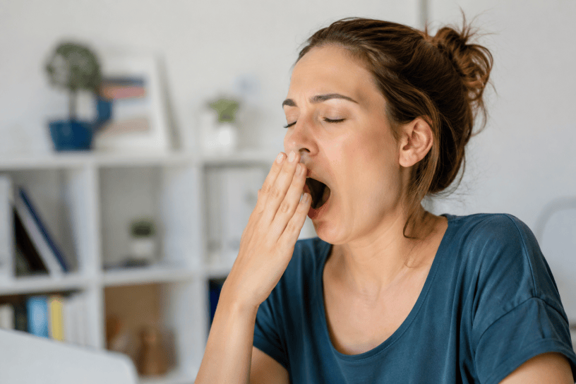 Giovane donna che sbadiglia seduta in casa durante una pausa, immagine che rappresenta la stanchezza, il sonno e i meccanismi naturali del corpo umano.