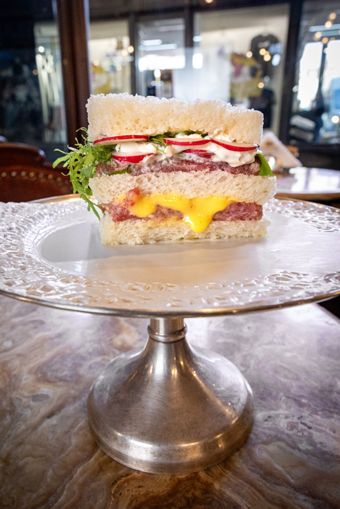 A layered sandwich on a decorative cake stand, featuring white bread, sliced radishes, leafy greens, mayonnaise, and melted cheese.