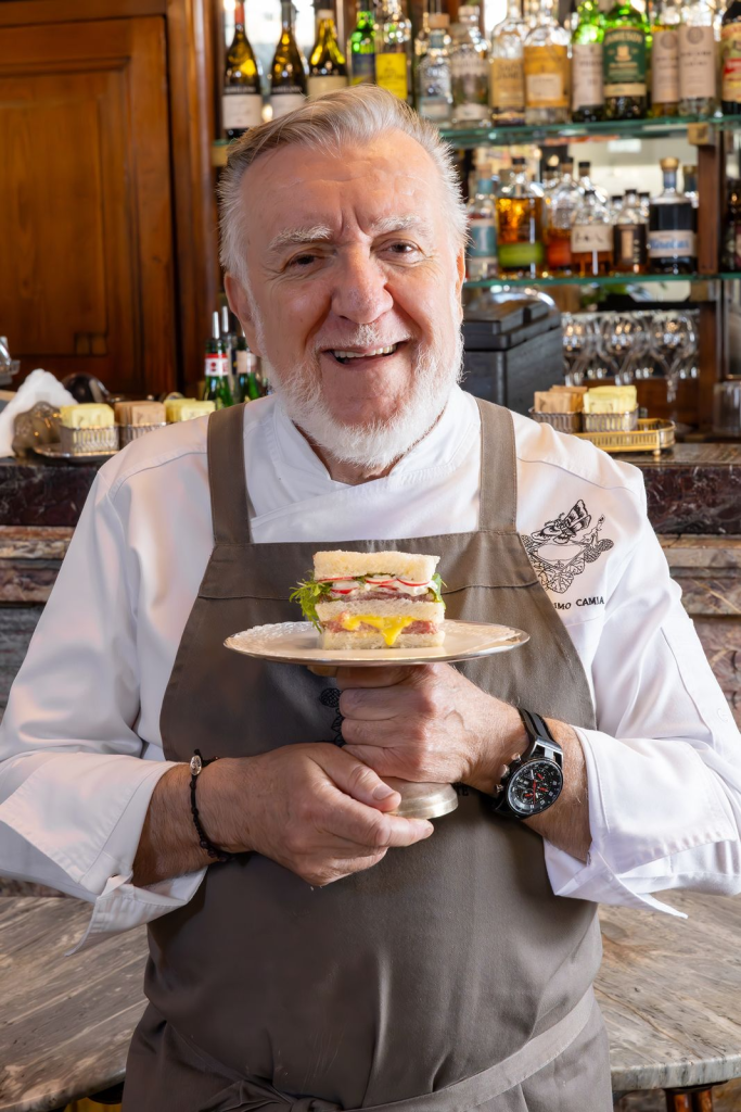 A smiling chef holding a plate with a sandwich in a well-stocked bar setting.