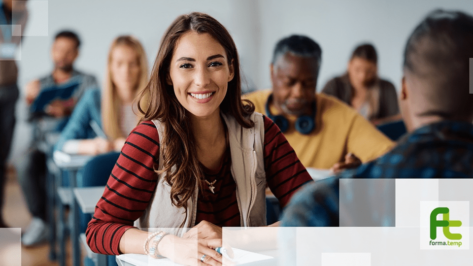 A smiling young woman sitting at a desk in a classroom, with fellow students in the background. The woman has long brown hair and is wearing a striped shirt and a white vest. The setting appears to be an educational environment.