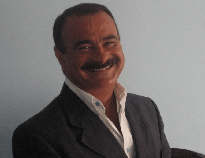 Smiling man wearing a black suit jacket over a white shirt, with a light blue background.