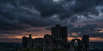 Person flying a colorful kite near dark ruins of a castle at sunset.