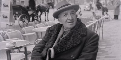 Ritratto in bianco e nero del poeta e scrittore italiano Vincenzo Cardarelli seduto a un tavolino all'aperto in una piazza italiana, fotografato da Paolo Monti nel 1957.