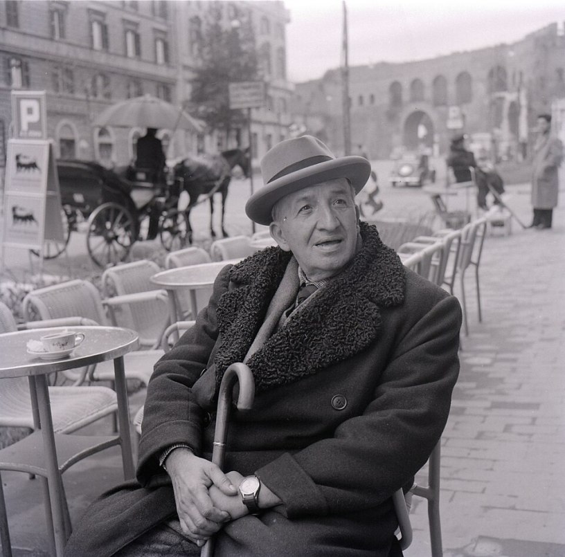 Ritratto in bianco e nero del poeta e scrittore italiano Vincenzo Cardarelli seduto a un tavolino all'aperto in una piazza italiana, fotografato da Paolo Monti nel 1957.