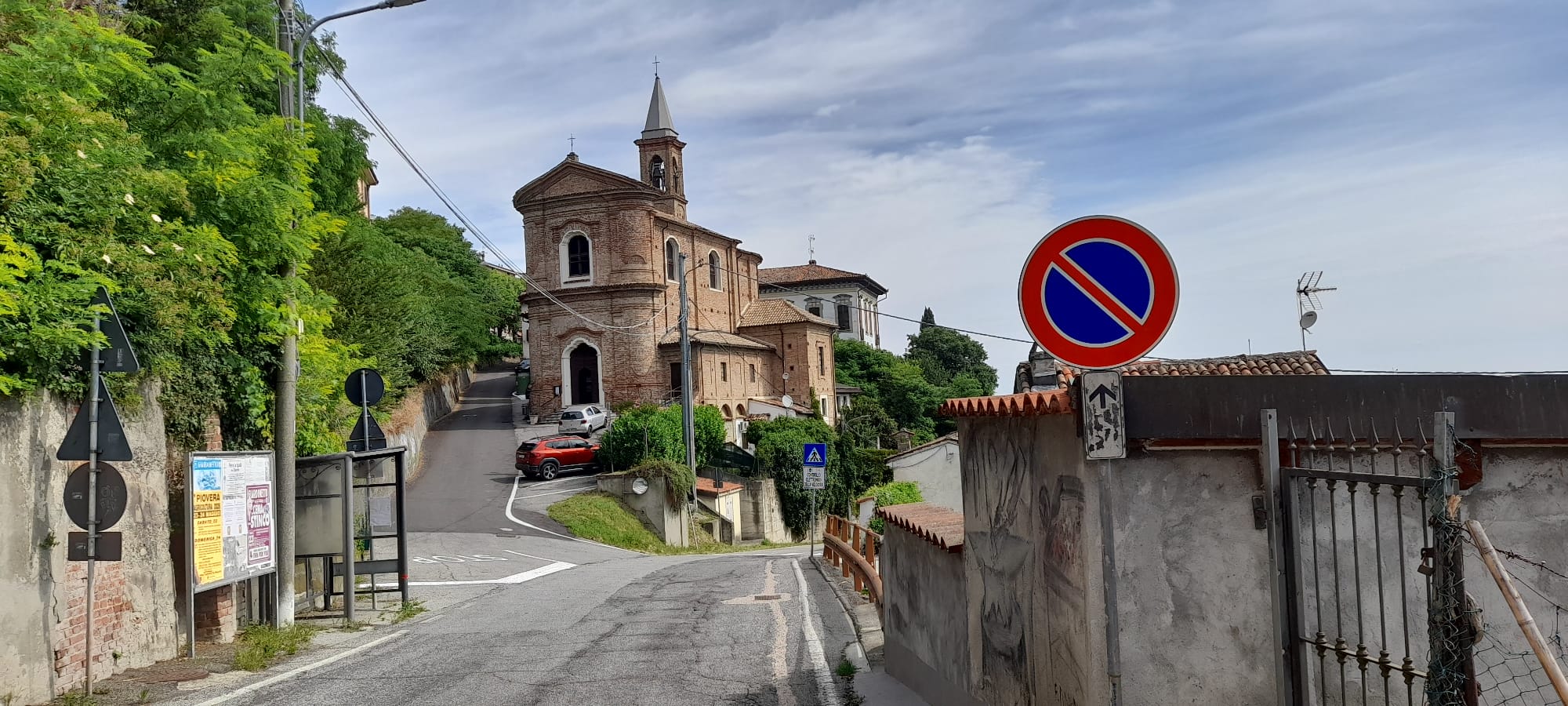 Vista del centro storico di Pecetto di Valenza con la chiesa parrocchiale in mattoni, la strada che sale verso il borgo e il paesaggio collinare del Monferrato in una giornata estiva.