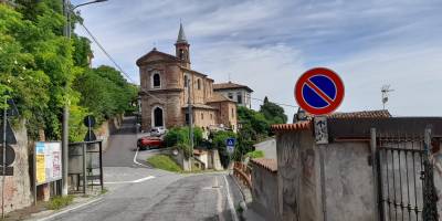 Vista del centro storico di Pecetto di Valenza con la chiesa parrocchiale in mattoni, la strada che sale verso il borgo e il paesaggio collinare del Monferrato in una giornata estiva.