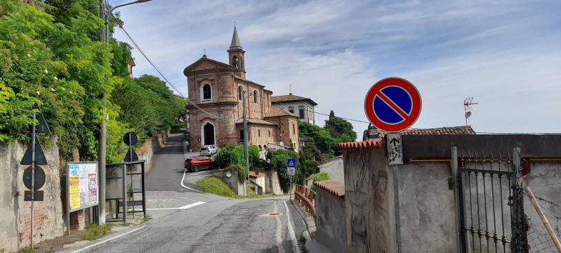 Vista del centro storico di Pecetto di Valenza con la chiesa parrocchiale in mattoni, la strada che sale verso il borgo e il paesaggio collinare del Monferrato in una giornata estiva.