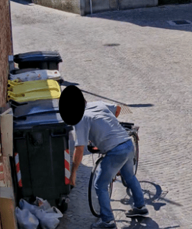 Person on a bicycle rummaging through a trash bin on a cobblestone street.