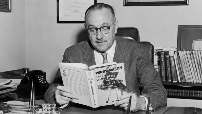 Ritratto fotografico in bianco e nero di Erle Stanley Gardner seduto alla scrivania mentre legge un romanzo di Perry Mason, circondato da libri, documenti e oggetti d’epoca.
