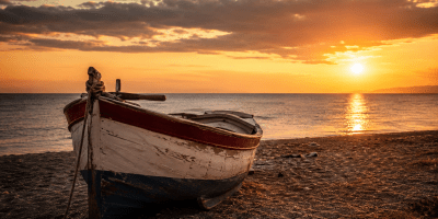 Barca in legno lasciata sulla spiaggia al tramonto, con il mare calmo e il cielo dorato sullo sfondo, immagine evocativa del tempo, della memoria e della nostalgia.
