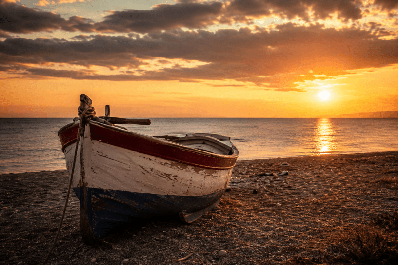 Barca in legno lasciata sulla spiaggia al tramonto, con il mare calmo e il cielo dorato sullo sfondo, immagine evocativa del tempo, della memoria e della nostalgia.