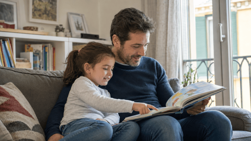 Padre seduto sul divano mentre legge un albo illustrato alla figlia, che indica con curiosità una figura sulla pagina.