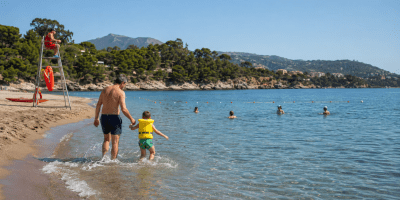 Un padre accompagna in mare il proprio bambino con il giubbotto salvagente, mentre un bagnino sorveglia la spiaggia.