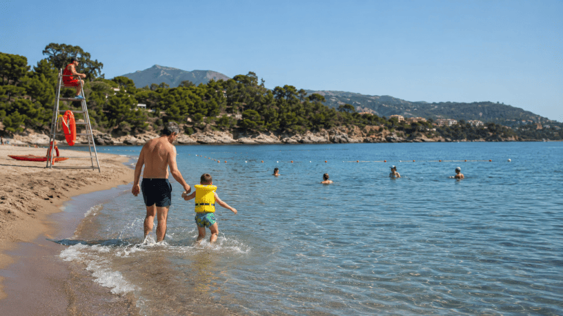 Un padre accompagna in mare il proprio bambino con il giubbotto salvagente, mentre un bagnino sorveglia la spiaggia.