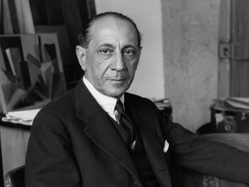 Ritratto fotografico in bianco e nero di Giacomo Balla nel suo studio, seduto accanto a tele e materiali artistici.