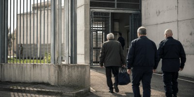 Uomo anziano accompagnato da alcuni agenti verso l’ingresso di un istituto penitenziario, immagine simbolica del caso Mario Roggero e dell’attesa per la possibile grazia.