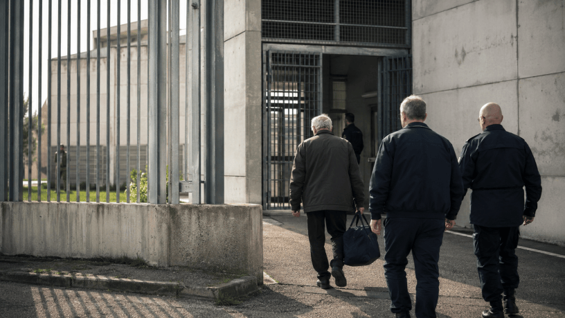 Uomo anziano accompagnato da alcuni agenti verso l’ingresso di un istituto penitenziario, immagine simbolica del caso Mario Roggero e dell’attesa per la possibile grazia.
