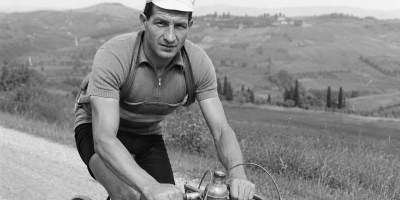 Gino Bartali in bicicletta su una strada di campagna, ritratto in bianco e nero durante un allenamento tra le colline toscane.