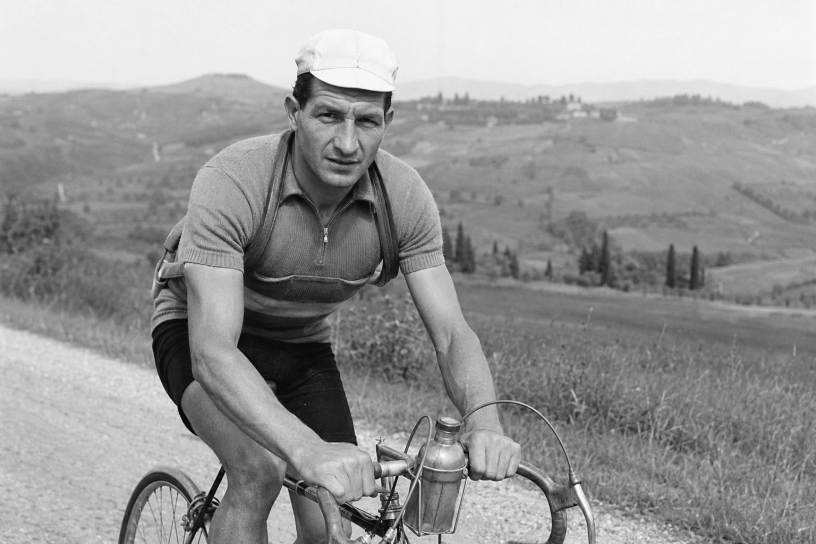 Gino Bartali in bicicletta su una strada di campagna, ritratto in bianco e nero durante un allenamento tra le colline toscane.