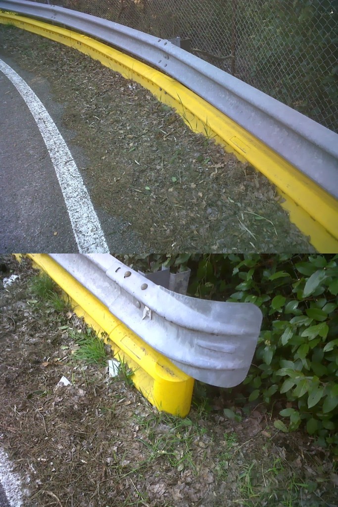 A close-up view of a curved highway guardrail, featuring a yellow bottom and a metallic gray top, with vegetation and gravel in the surrounding area.
