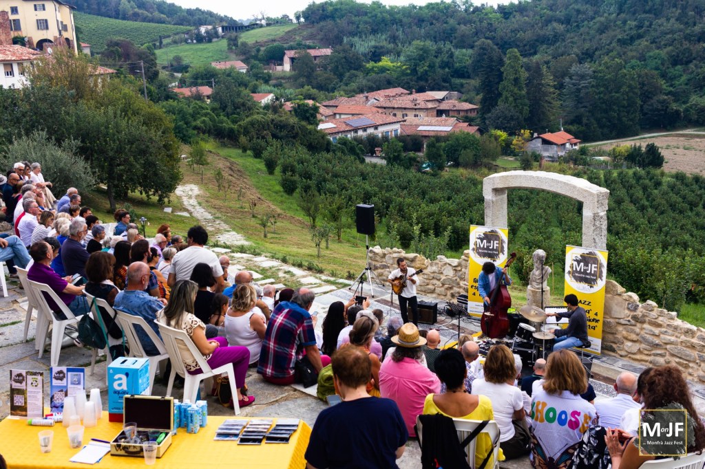A live jazz performance taking place outdoors, with musicians playing instruments in front of an audience seated on chairs. Scenic vineyards and houses in the background.