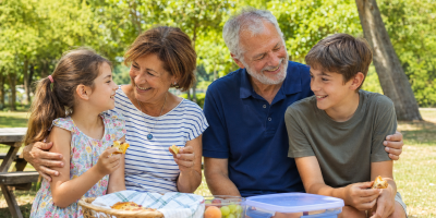 Due nonni trascorrono una giornata estiva al parco con i nipoti, condividendo la merenda e un momento di affetto familiare.