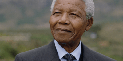 Ritratto fotografico realistico di Nelson Mandela in età avanzata, vestito in modo formale e ripreso all’aperto con espressione serena e riflessiva.