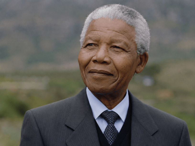 Ritratto fotografico realistico di Nelson Mandela in età avanzata, vestito in modo formale e ripreso all’aperto con espressione serena e riflessiva.