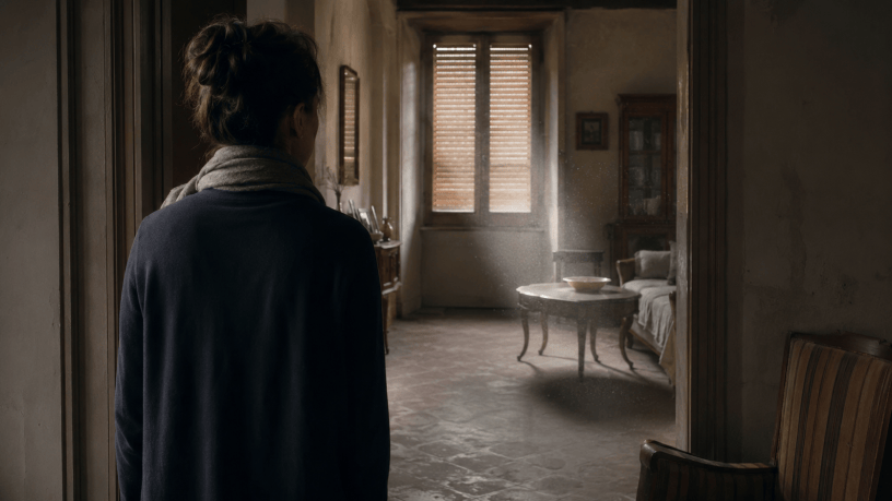 Donna vista di spalle si ferma sulla soglia di una stanza silenziosa, con mobili immobili, persiane chiuse e polvere sospesa nella luce.