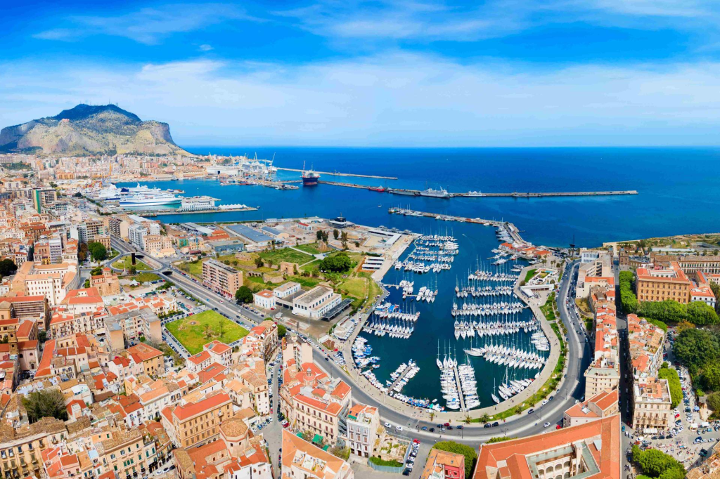 Aerial view of a coastal city with a harbor filled with yachts and boats, surrounded by buildings and green spaces, under a clear blue sky.