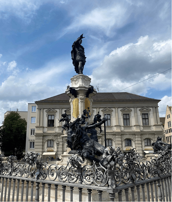 A large statue of a historical figure on a tall pedestal, surrounded by intricate bronze sculptures and a decorative wrought iron fence, with a building in the background and a cloudy sky above.