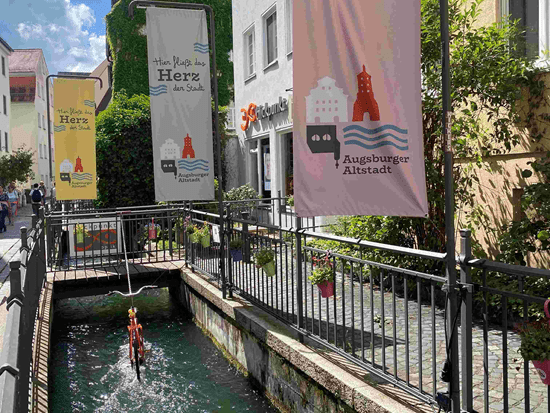A picturesque urban scene featuring decorative banners along a narrow canal in Augsburg's Old Town, with a bicycle hanging above the water.