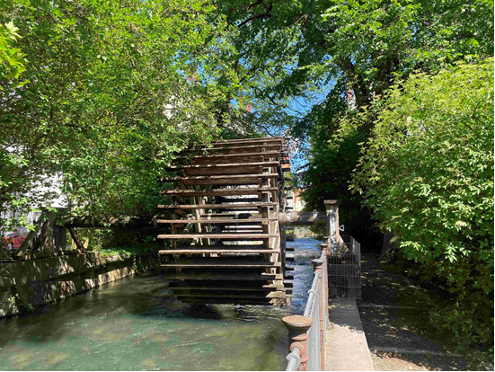 A wooden waterwheel situated over a stream, surrounded by lush green foliage and trees, with a blue sky visible in the background.