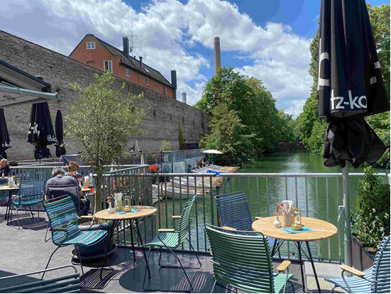 Outdoor cafe by a river with tables and chairs, surrounded by trees and a historic stone wall, under a partly cloudy sky.