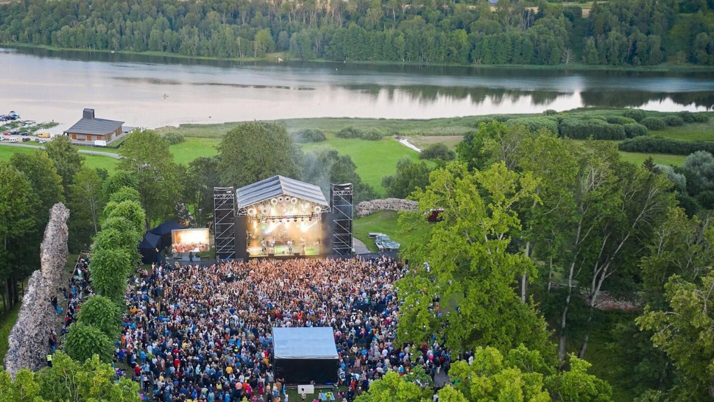 Aerial view of a large outdoor concert with a crowd gathered in front of a brightly lit stage, surrounded by trees and a serene lake in the background.