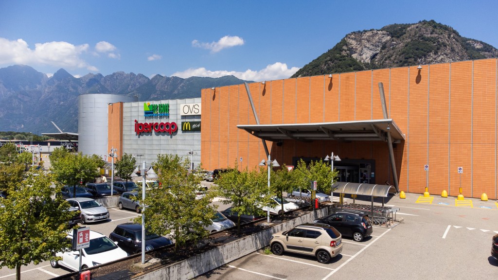 Exterior view of a shopping mall with the Ipercoop and OVS stores, featuring a McDonald's sign, surrounded by parking and trees, against a backdrop of mountains and blue sky.