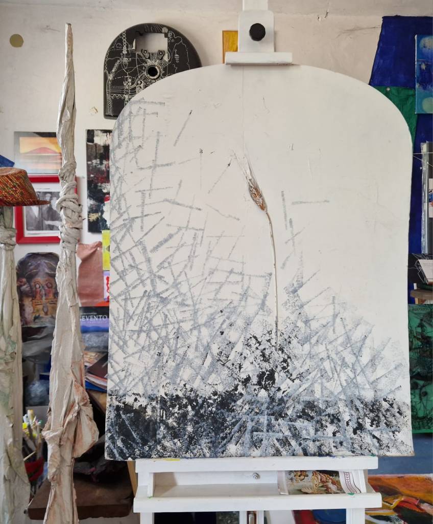 An abstract painting on an easel featuring a textured white background with dark, scratch-like patterns and a single grass-like element in the center, set in an art studio with various other artworks and materials visible in the background.
