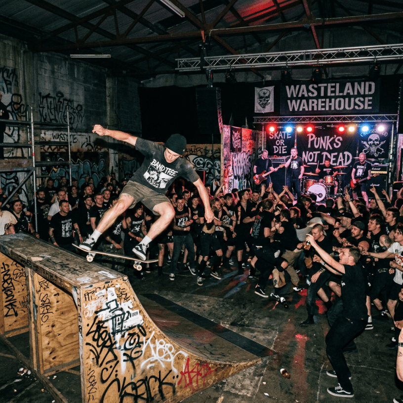 Skateboarder doing a trick on a graffiti-covered ramp at an indoor punk rock concert with a live band and enthusiastic crowd
