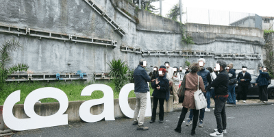 Visitatori riuniti davanti alle grandi lettere C.O.M. nel cortile delle case popolari del quartiere Caina a Carrara, sede delle nuove opere di arte contemporanea.