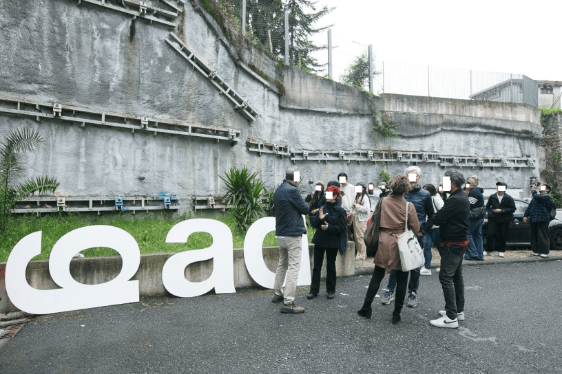 Visitatori riuniti davanti alle grandi lettere C.O.M. nel cortile delle case popolari del quartiere Caina a Carrara, sede delle nuove opere di arte contemporanea.