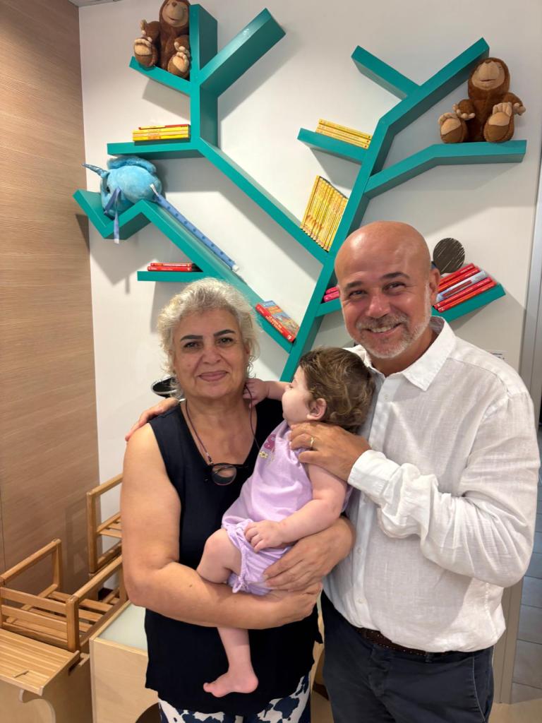 A woman and a man are smiling and holding a young child in a playful indoor environment. Behind them, there is a colorful bookshelf in the shape of a tree, decorated with toys and children's books.