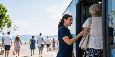 Una lavoratrice dei trasporti aiuta una donna anziana a salire su un autobus, mentre turisti camminano sul lungomare in una luminosa giornata estiva.