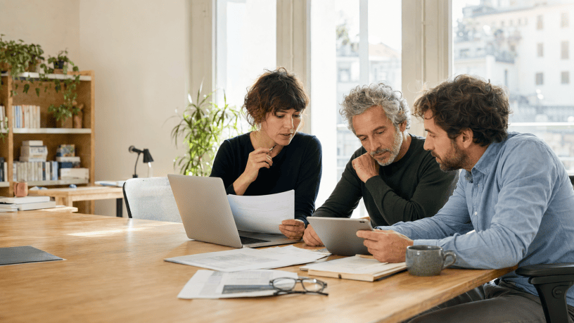 Tre collaboratori di una redazione digitale confrontano documenti, un computer portatile e un tablet attorno a un tavolo di lavoro.